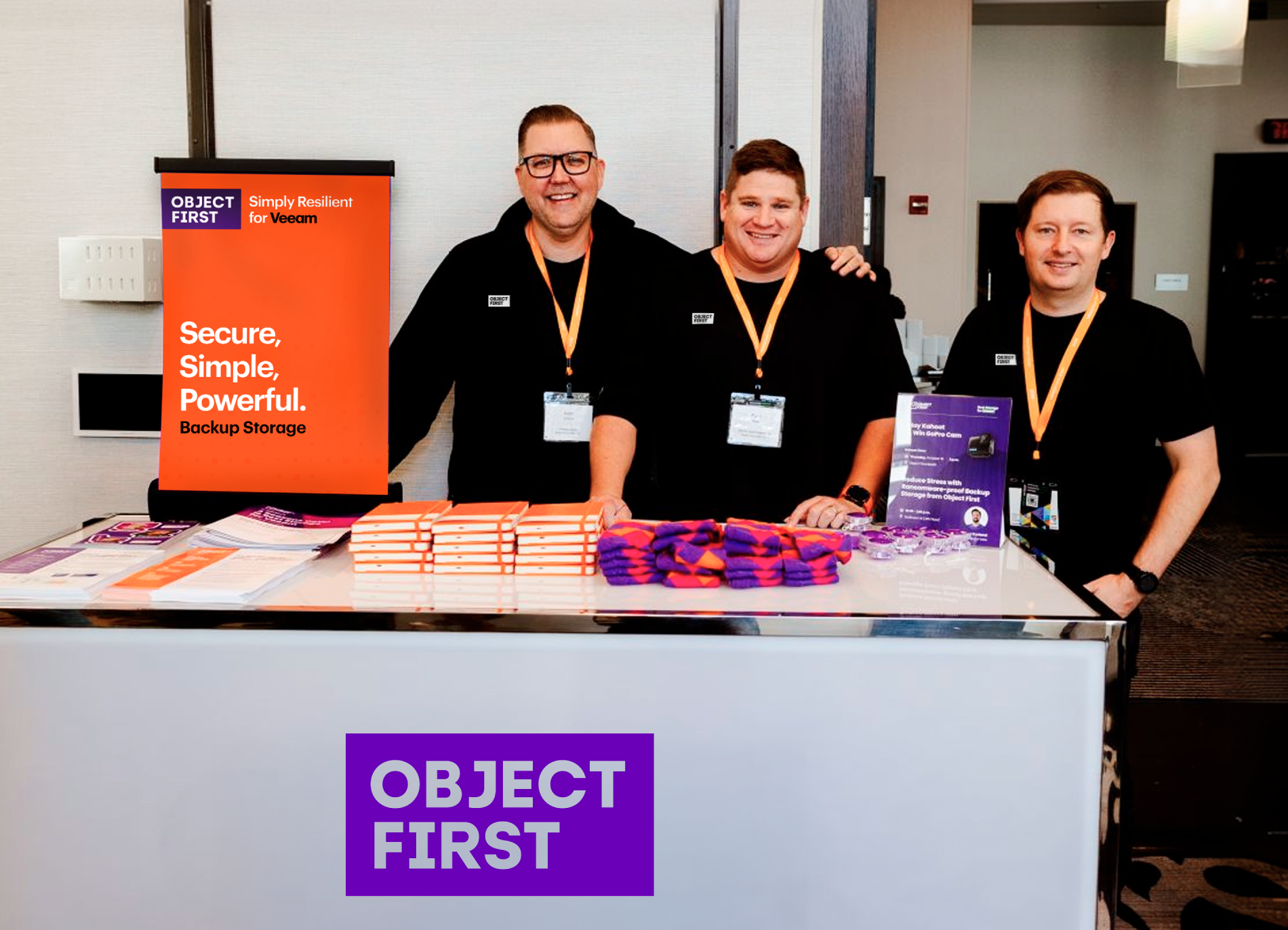 An event booth for Object First, staffed by three smiling men in black t-shirts and orange lanyards. A large orange banner to the left reads, 'OBJECT FIRST. Simply Resilient for Veeam. Secure, Simple, Powerful. Backup Storage.' The counter in front displays stacks of orange notebooks, neatly folded piles of orange and purple socks, and promotional flyers. The front of the booth features a prominent white 'OBJECT FIRST' logo in a purple box.