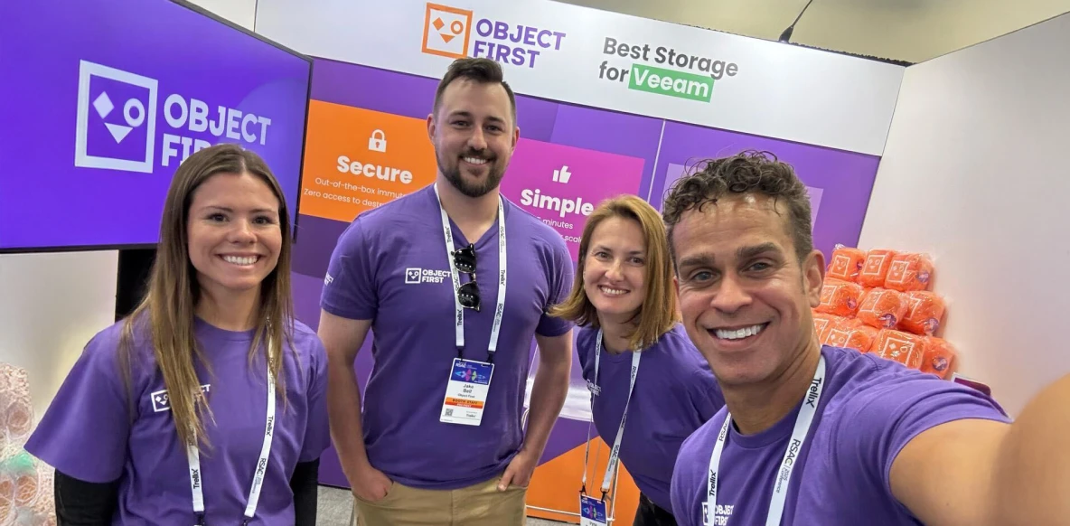 A group of seven people posing together in a brightly lit office or conference setting, with four women wearing matching purple shirts, standing in front of a purple banner that reads "OBJECT FIRST" and promotes storage solutions for Veeam.