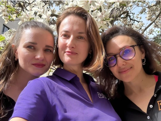 Three women outdoors under a blooming tree, smiling at the camera, with one of them wearing purple sunglasses.
